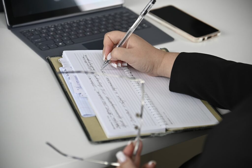 Uma Pessoa Escrevendo Em Um Caderno Com Um Laptop Ao Fundo 0KT9gxj_BdM