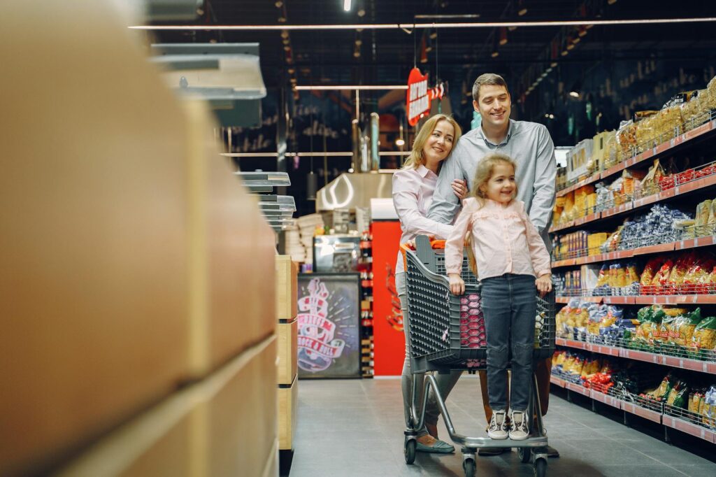 Family enjoying a shopping trip in a supermarket, with a child in a cart and parents smiling.