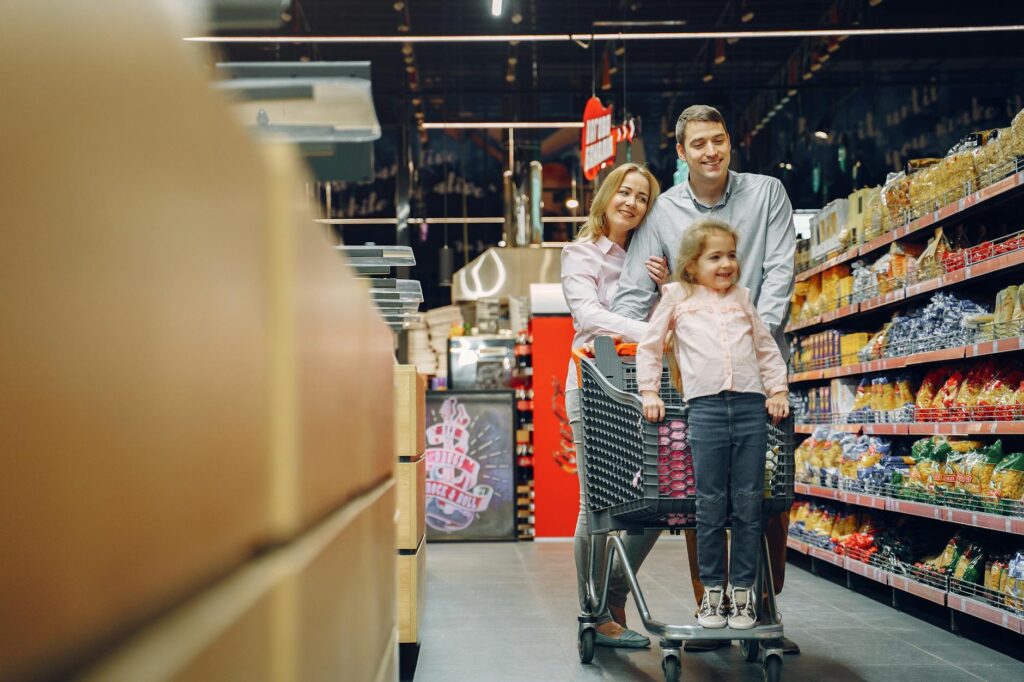 Family enjoying a shopping trip in a supermarket, with a child in a cart and parents smiling.